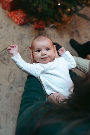 Baby in white sweater lying on green surface raises arms joyfully during holiday season.