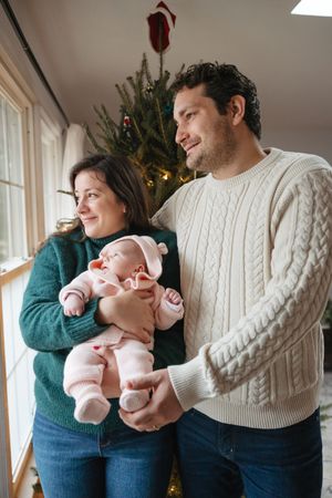 A family shares a tender moment by their Christmas tree while holding their newborn baby in winter sweaters.