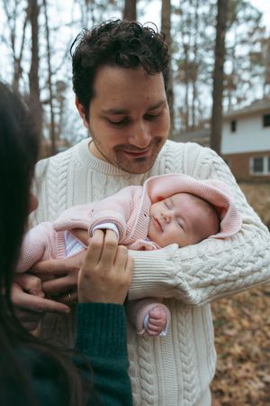 A tender moment between a parent wearing a white cable knit sweater and a sleeping baby in pink clothing outdoors.
