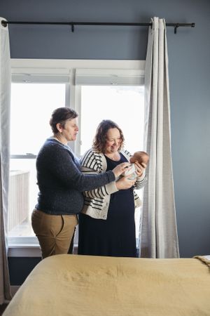 Two people stand by a window holding and gazing at a baby in a cozy bedroom setting with natural light.