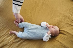 A baby in a soft blue sleeper outfit lies on a golden-colored blanket while someone's hand touches their arm.