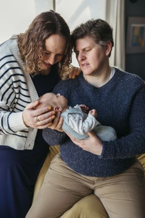 A couple in sweaters tenderly holds their newborn baby while sitting together near a window.