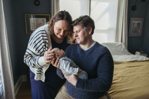 A couple in casual sweaters tenderly holds and gazes at their newborn baby by a sunlit window.