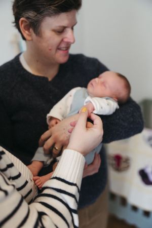 A tiny newborn baby in white sleepwear being cradled while someone in a striped sweater holds their tiny hand.