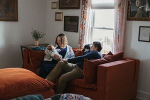 Two people relax together on a burnt orange couch near a window in a cozy living room with framed artwork on walls.