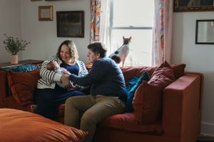 Two people relax together on a brown couch with a cat in a sunlit living room by a window.