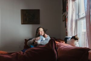Person relaxes on red couch by window with cat while wearing striped sweater in cozy room setting.