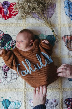 Small baby wearing brown knit sweater with embroidered text lies on colorful patchwork quilt while hands reach from sides.