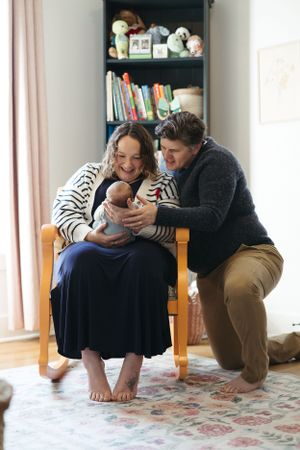 A couple sits together on a wooden chair in a cozy nursery corner with bookshelves and stuffed animals in the background.