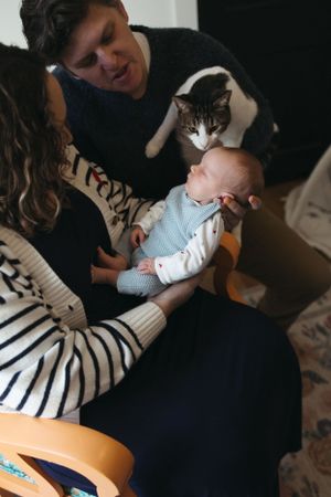 Family sits together on couch with black and white cat curiously watching over small baby in blue outfit.