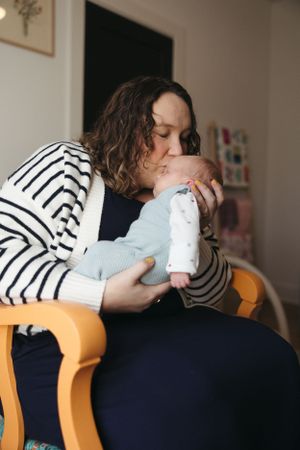 A person in a striped sweater sits in an orange chair, tenderly kissing an infant wrapped in light blue clothing.