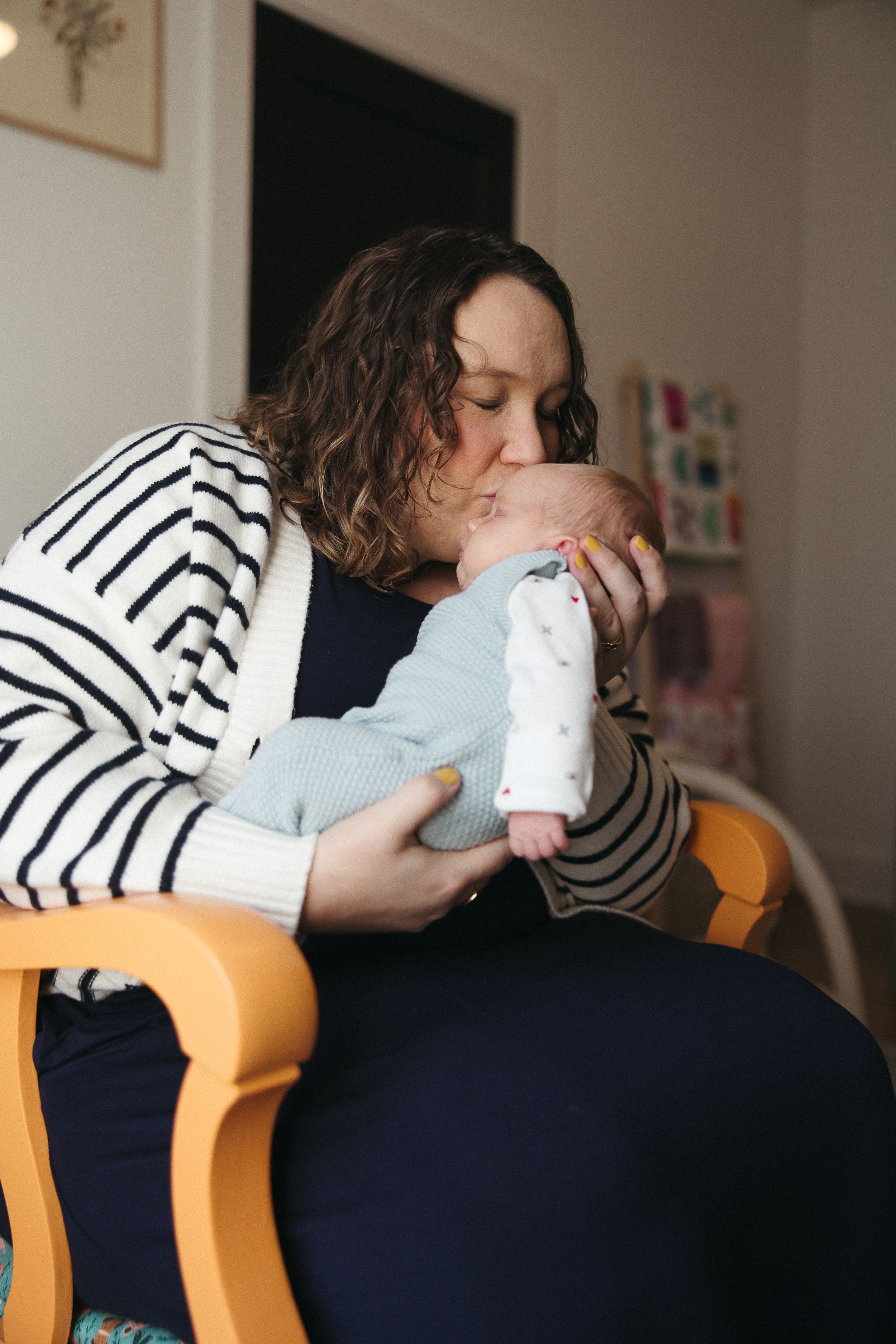 A person in a striped sweater sits in an orange chair, tenderly kissing an infant wrapped in light blue clothing.