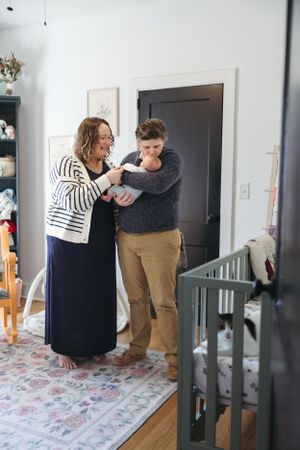 Two people stand together in a cozy living room with a patterned rug, sharing a tender moment by a dark doorway.