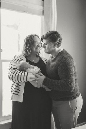 A tender moment captured in black and white as two people share an intimate moment while holding a newborn by a window.