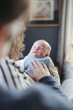 A peaceful sleeping newborn baby in a light blue sweater is held gently by caring hands in a cozy indoor setting.