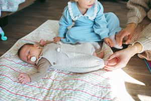 A baby wearing blue and gray stripes lies on a colorful patterned blanket while hands reach to comfort them.