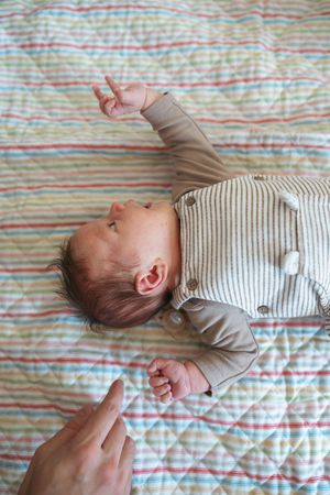 A baby in a striped onesie lies on a colorful striped blanket while someone gently holds their tiny hand.
