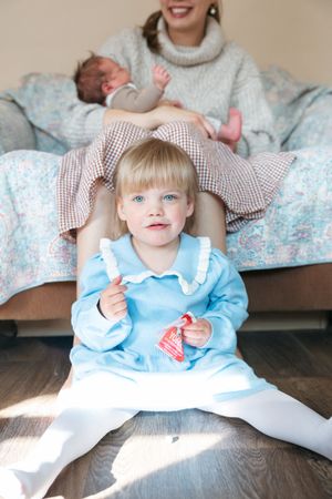 A toddler in a blue dress sits on a floral patterned surface while an infant rests behind on a couch.