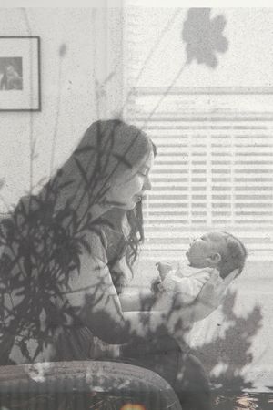 A tender black and white moment between parent and infant by a window with venetian blinds and leaf shadows.