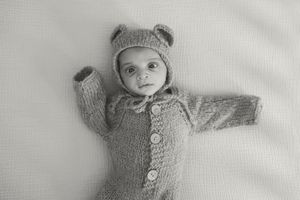A small baby wearing a gray knit beanie and sweater lies on a white blanket.
