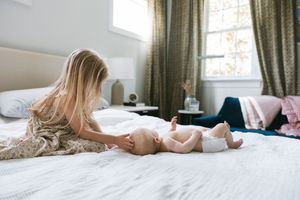 Young girl with long blonde hair playing with a baby in a diaper on a white bed in a bright bedroom.