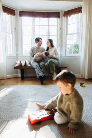 A family relaxes together in a sunny bay window while a child plays with toys on the floor.
