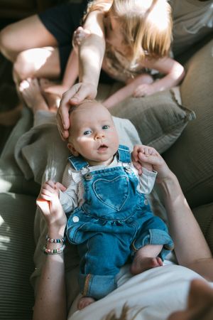 Baby in denim overalls with heart detail lying on adult lap while blonde child gently touches infant's head on sofa.