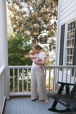 A mother holds her newborn baby on a white-railed porch beside a magnolia tree and black rocking chair.