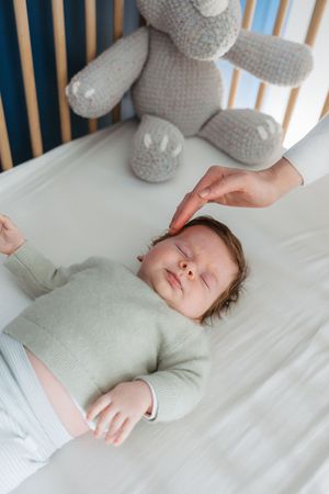 Sleeping baby in gray clothing lies in crib next to plush teddy bear.