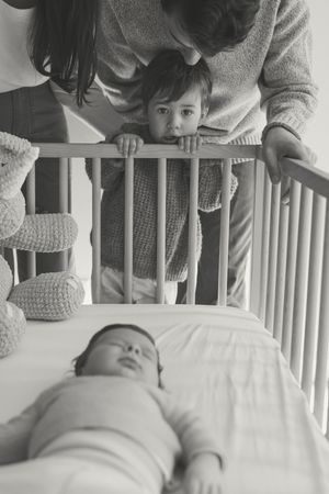A parent leans over a white crib to interact with their baby in a series of sweet black and white photographs.