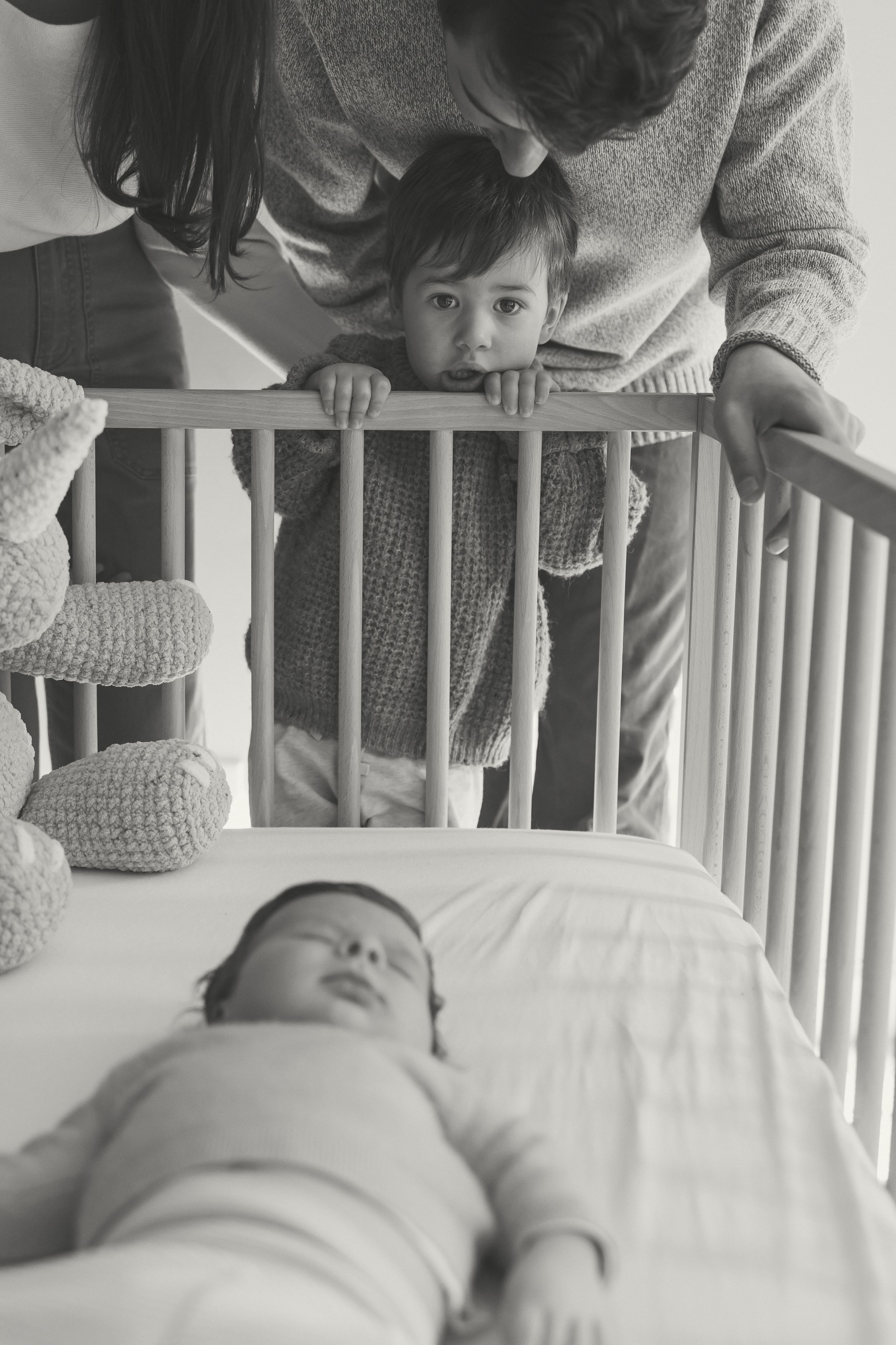 A parent leans over a white crib to interact with their baby in a series of sweet black and white photographs.