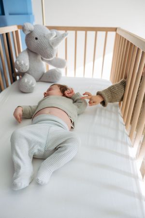 Baby sleeping peacefully in white crib with soft gray stuffed animal toys surrounding them on white bedding.