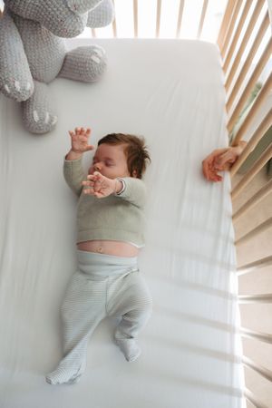 Baby sleeping peacefully in a white crib wearing striped pajamas with a plush toy nearby.