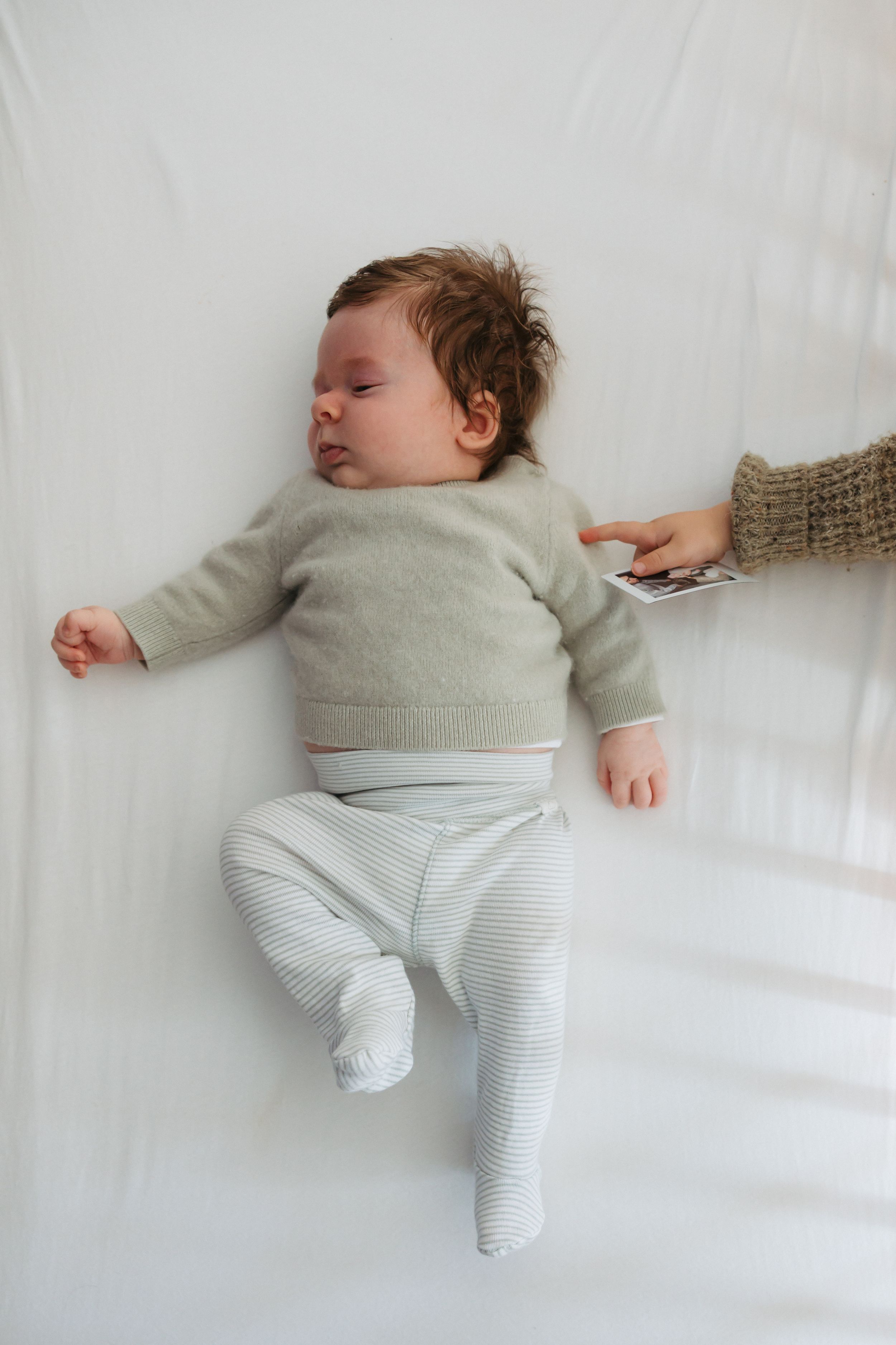 Baby wearing light gray sweater and striped pants sleeping peacefully on white bedding.