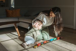 A child plays with a line of colorful toy trains on a light carpet in a home setting.