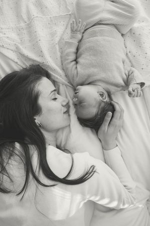 Tender black and white portrait of a mother and baby lying together on white bedding.