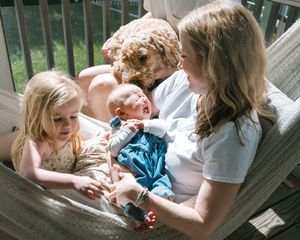 A mother sits in a hammock holding a newborn baby, with a young girl and a curly dog beside her.