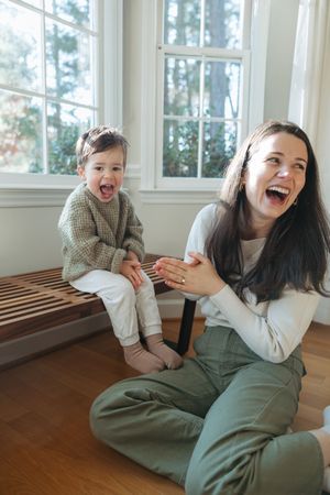 A parent and child share a joyful moment of laughter while sitting together by large windows in a sunlit room.