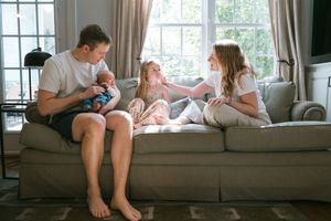 Family of four sitting on a cozy gray sofa, parents bonding with newborn baby and young daughter in bright living room.