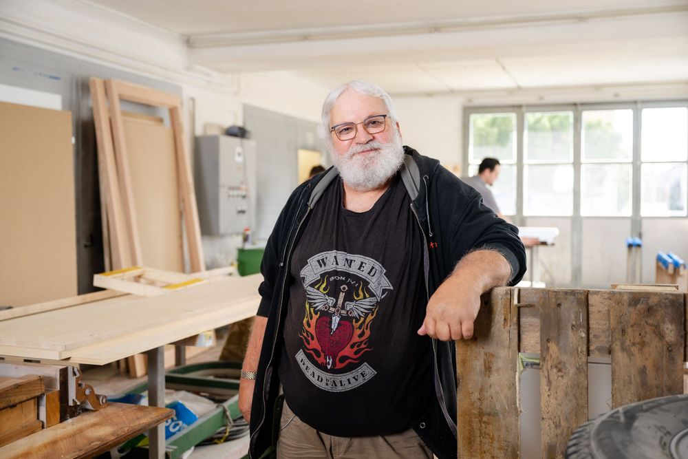 Workshop interior with person wearing black t-shirt and jacket standing near wooden furniture.