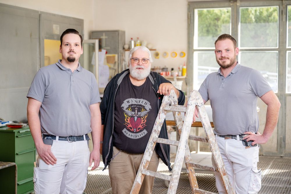 Three workers stand in a workshop environment near a ladder and storage shelves.