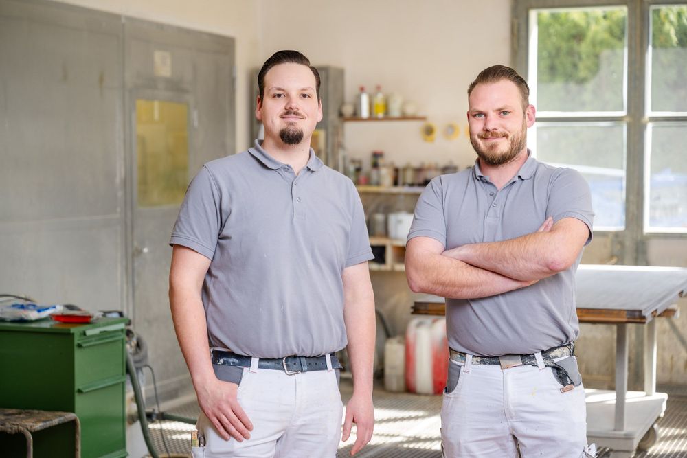 Two workers in matching grey polo shirts pose in an industrial workspace.
