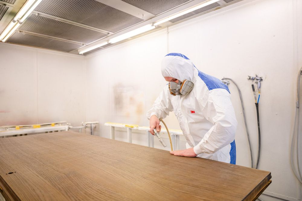 Person in protective white suit and mask spraying treatment on wooden surface in workshop.