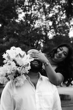 A woman playfully covers a man’s eyes from behind as he laughs, holding a large bouquet of fresh white lilies and chrysanthemums, set agains