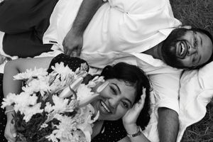 A joyful couple lies on a blanket in the grass, smiling up at the camera; the woman holds a bouquet of flowers while the man laughs heartily