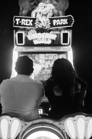 A rear-view shot of a couple at an arcade game; the woman turns back to smile at the camera, her arm gently around the man’s shoulder, glowi