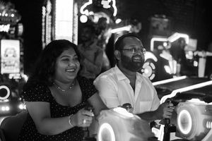 A close-up of a couple sitting side by side at an arcade game, both gripping joysticks, laughing with childlike excitement surrounded by glo