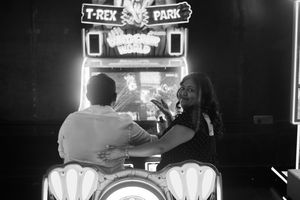 A couple sits side by side at an arcade game titled “T-Rex Park,” their backs to the camera, lost in the glowing screen, surrounded by nosta
