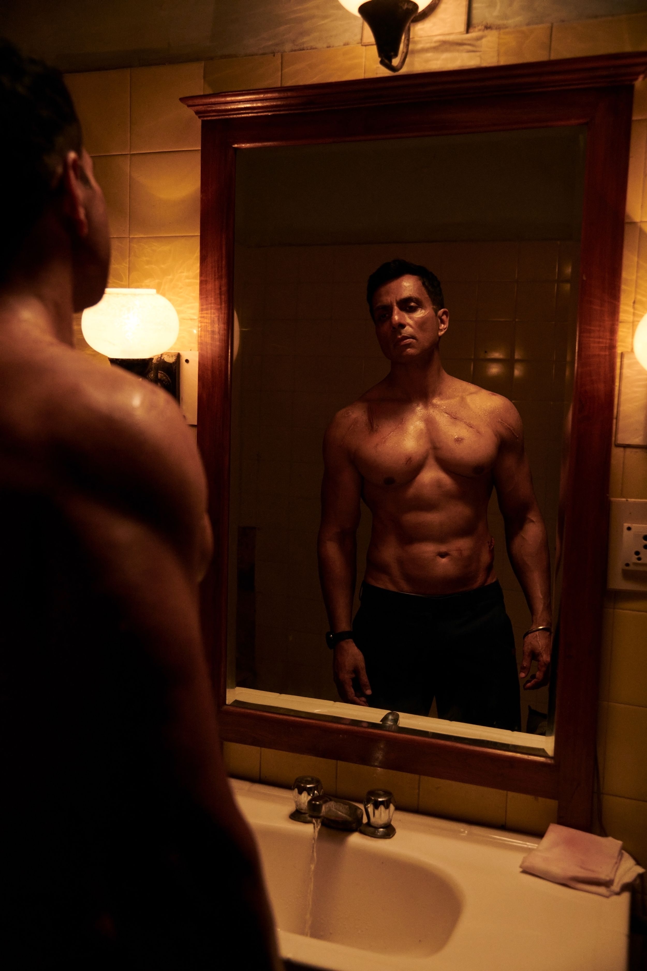 Man looking at himself in a bathroom mirror, shirtless, under warm lighting, reflecting a thoughtful and intense expression.