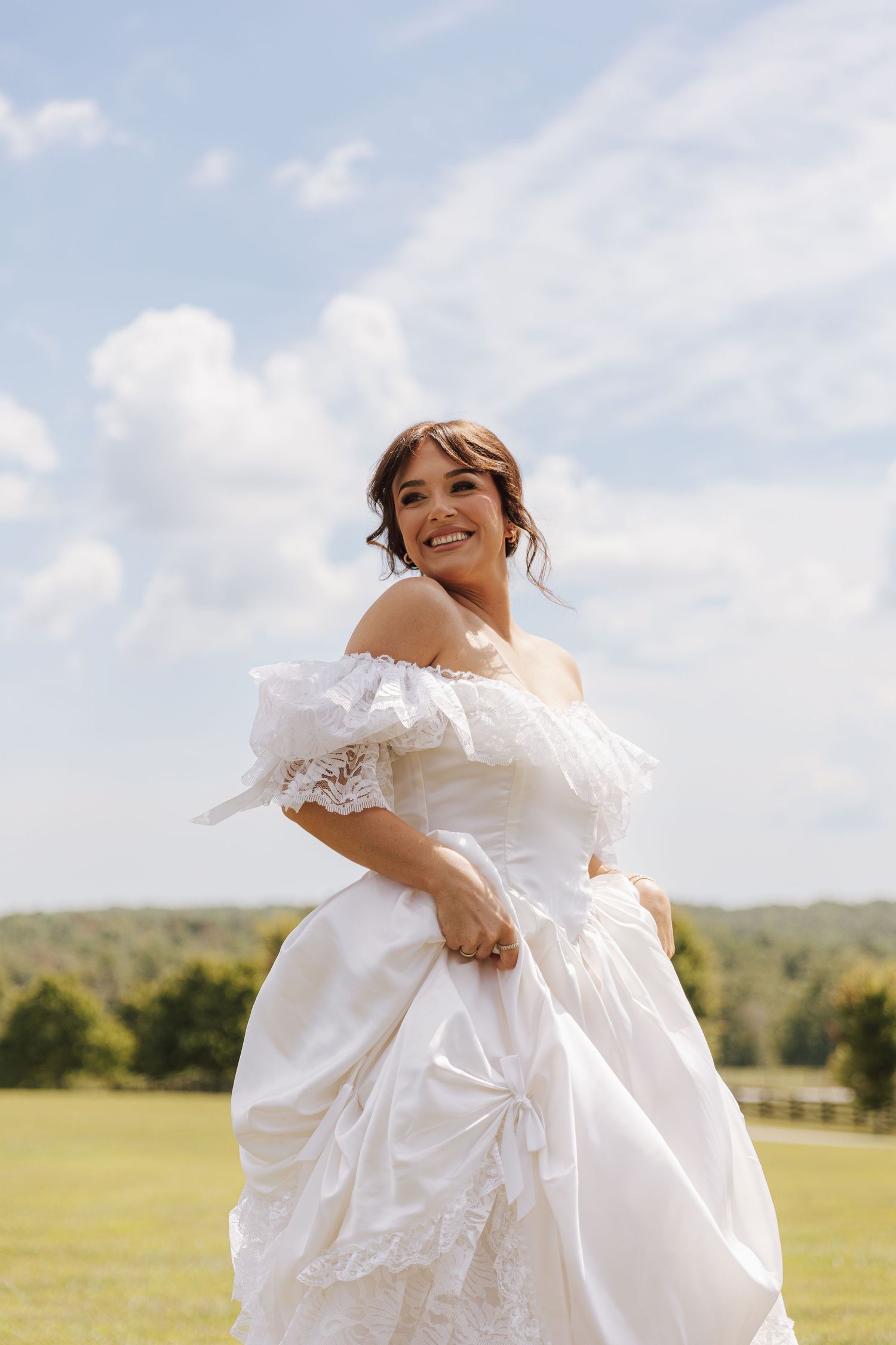 A bride wearing an elegant off-shoulder white wedding gown stands in a sunny field with a cloudy blue sky backdrop.